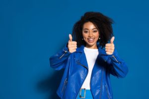smiling young woman holding two thumbs up on a blue background in a blue jacket oral health general dentistry dentist in Columbia South Carolina